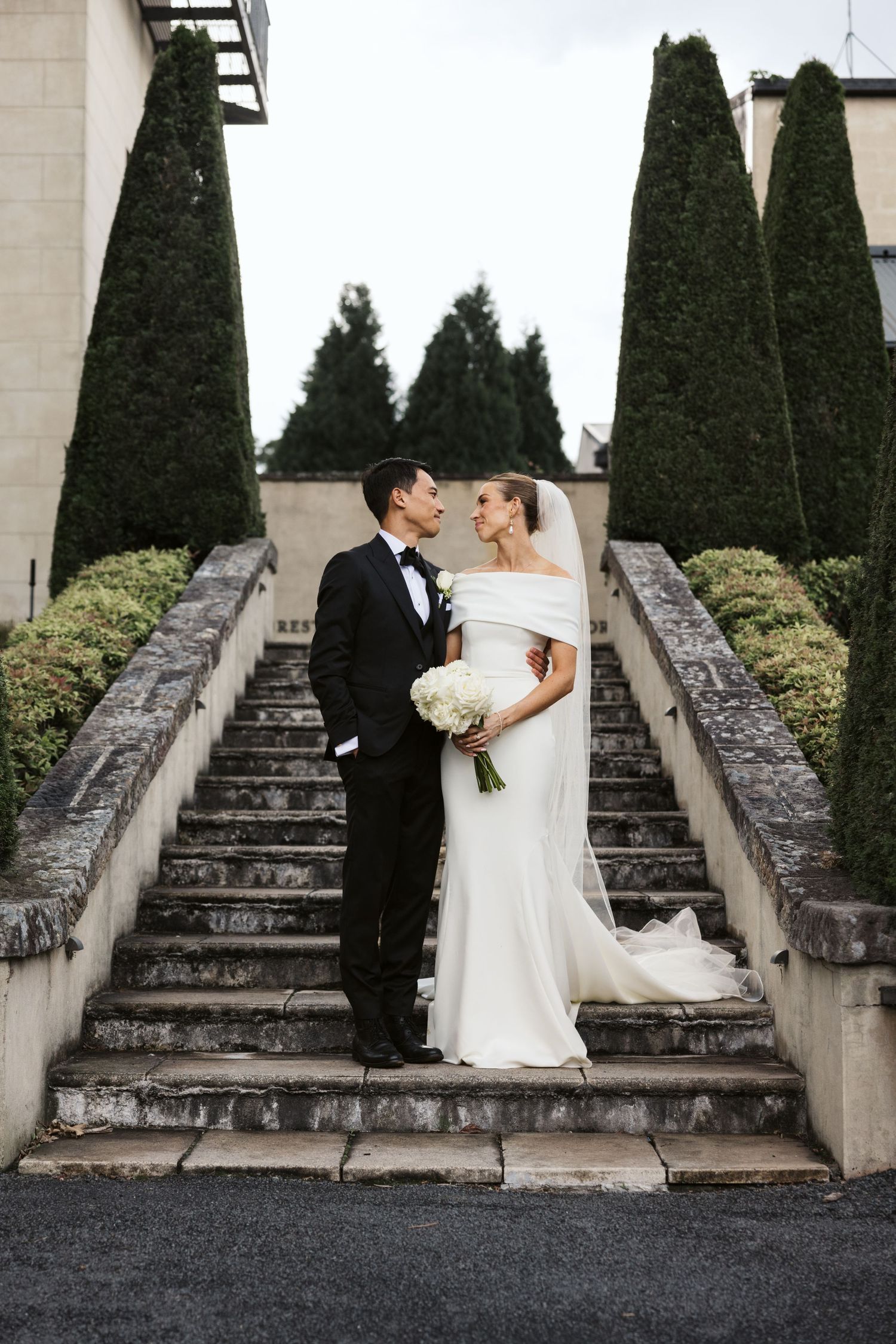 Elegant couple shares romantic moment on grand stone staircase flanked by manicured topiary trees at outdoor wedding venue.