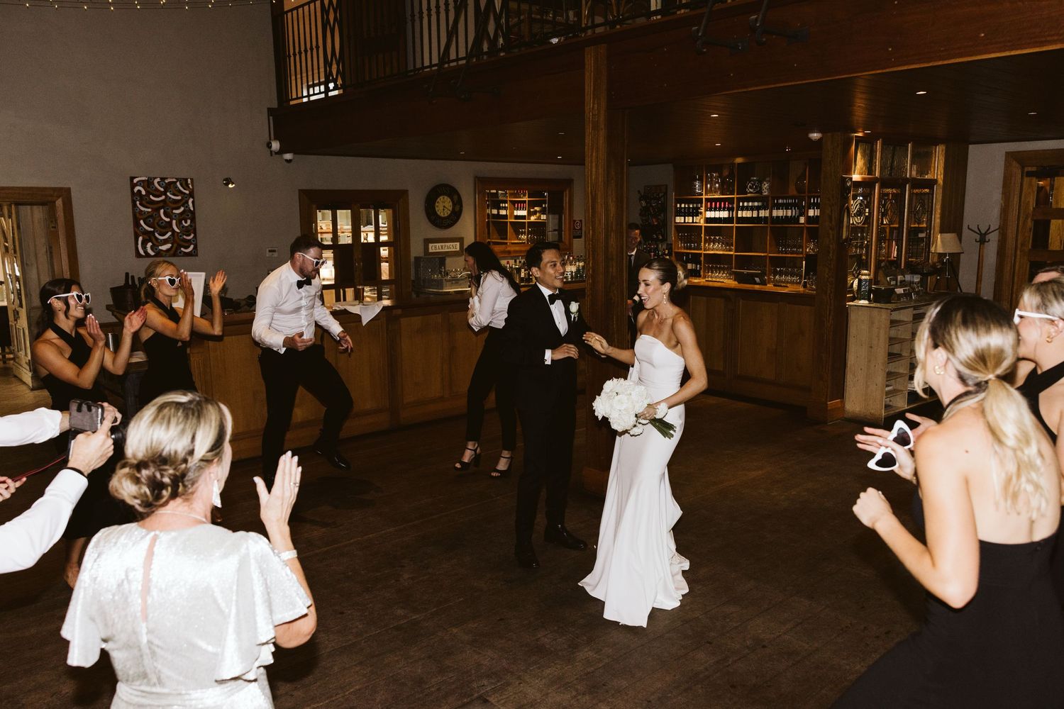 A couple dances at their wedding reception while guests take photos in a rustic venue with wooden walls.