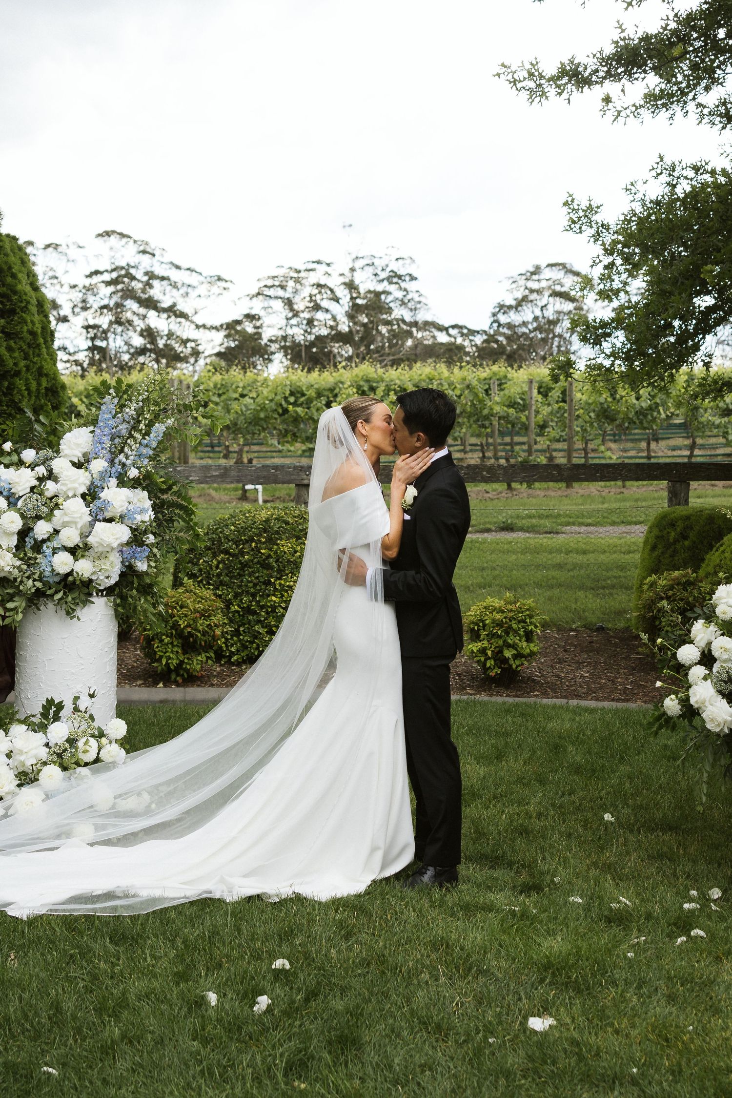 A wedding couple shares a romantic moment in a garden with white floral arrangements.