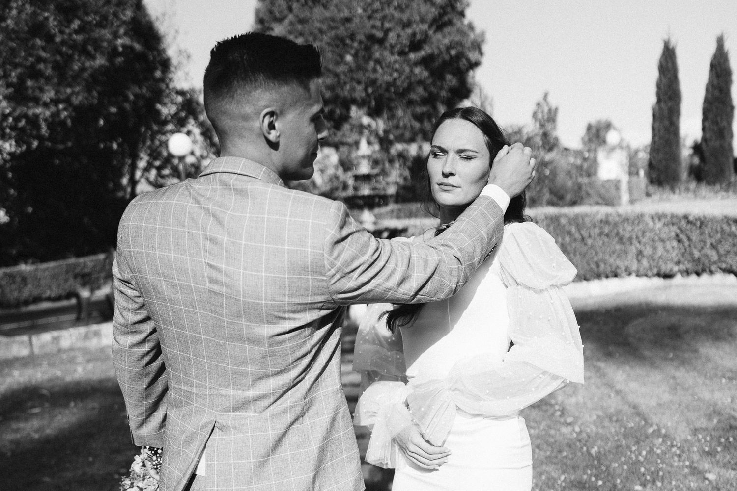 Black and white photo of a groom in plaid suit adjusting bride's hair outdoors at a garden wedding.