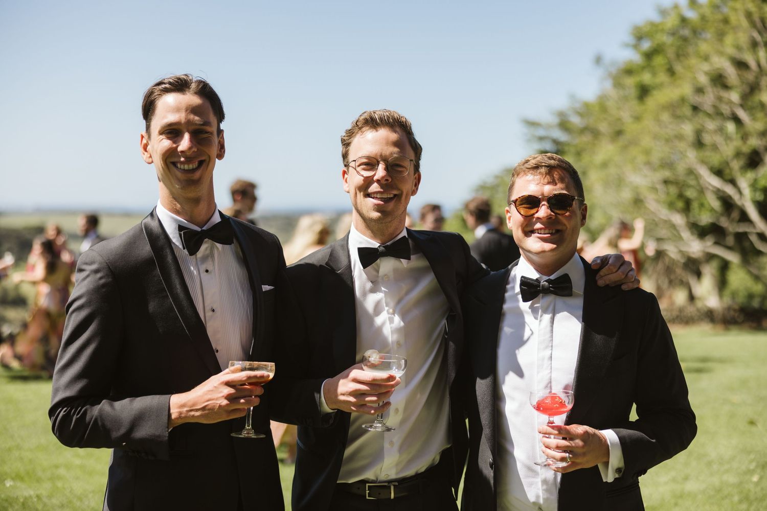 Three guests in formal attire enjoying drinks during outdoor celebration.