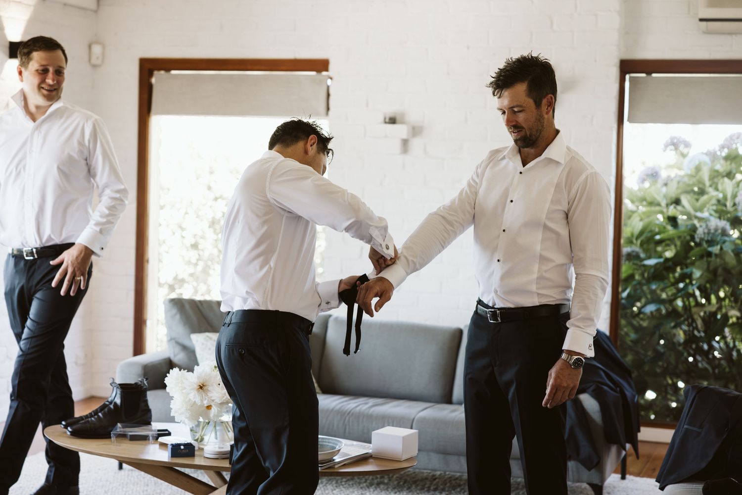 Wedding preparation scene showing groomsmen helping each other get dressed in a bright room.