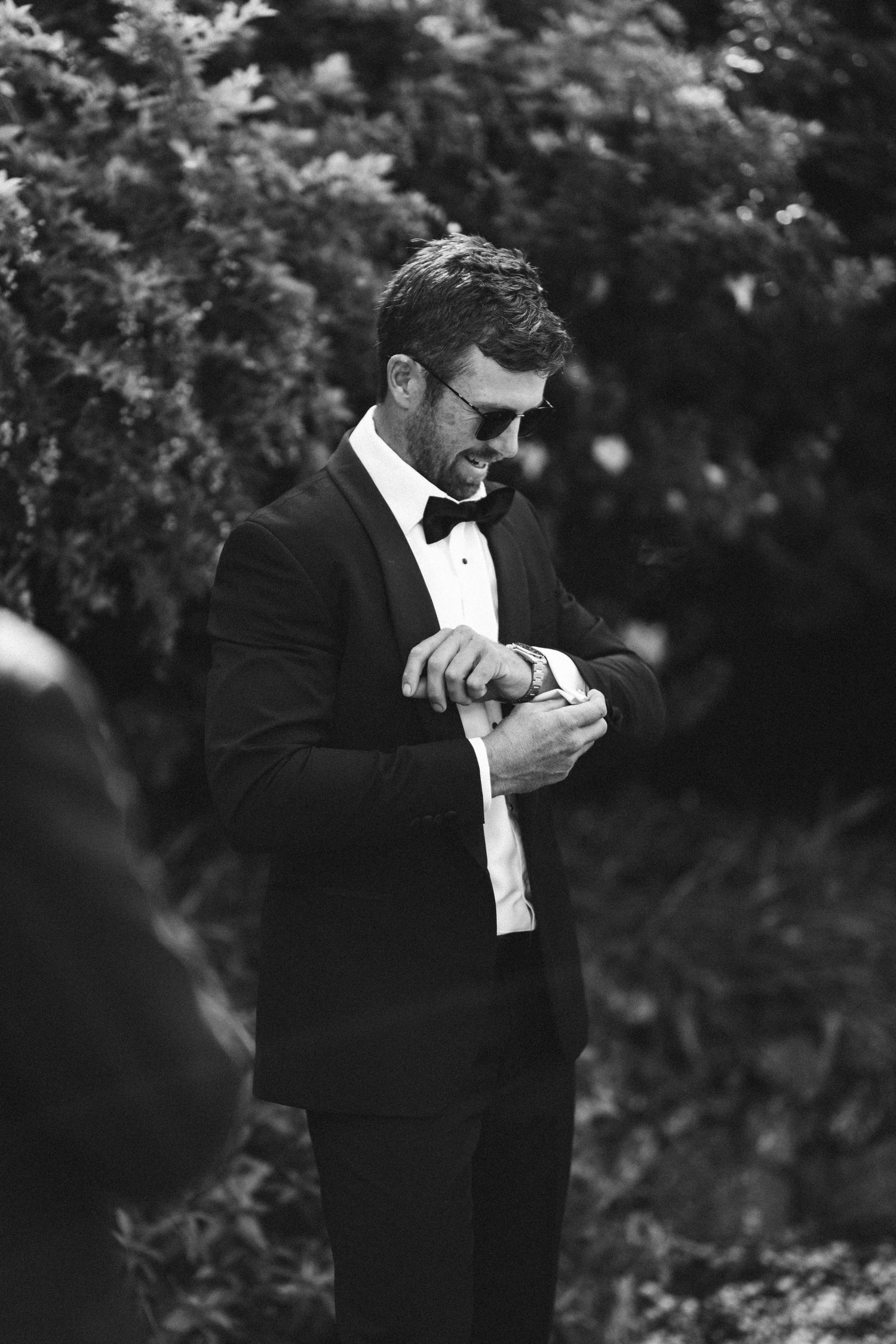 Person in black tuxedo and sunglasses checking watch outdoors in black and white photography.
