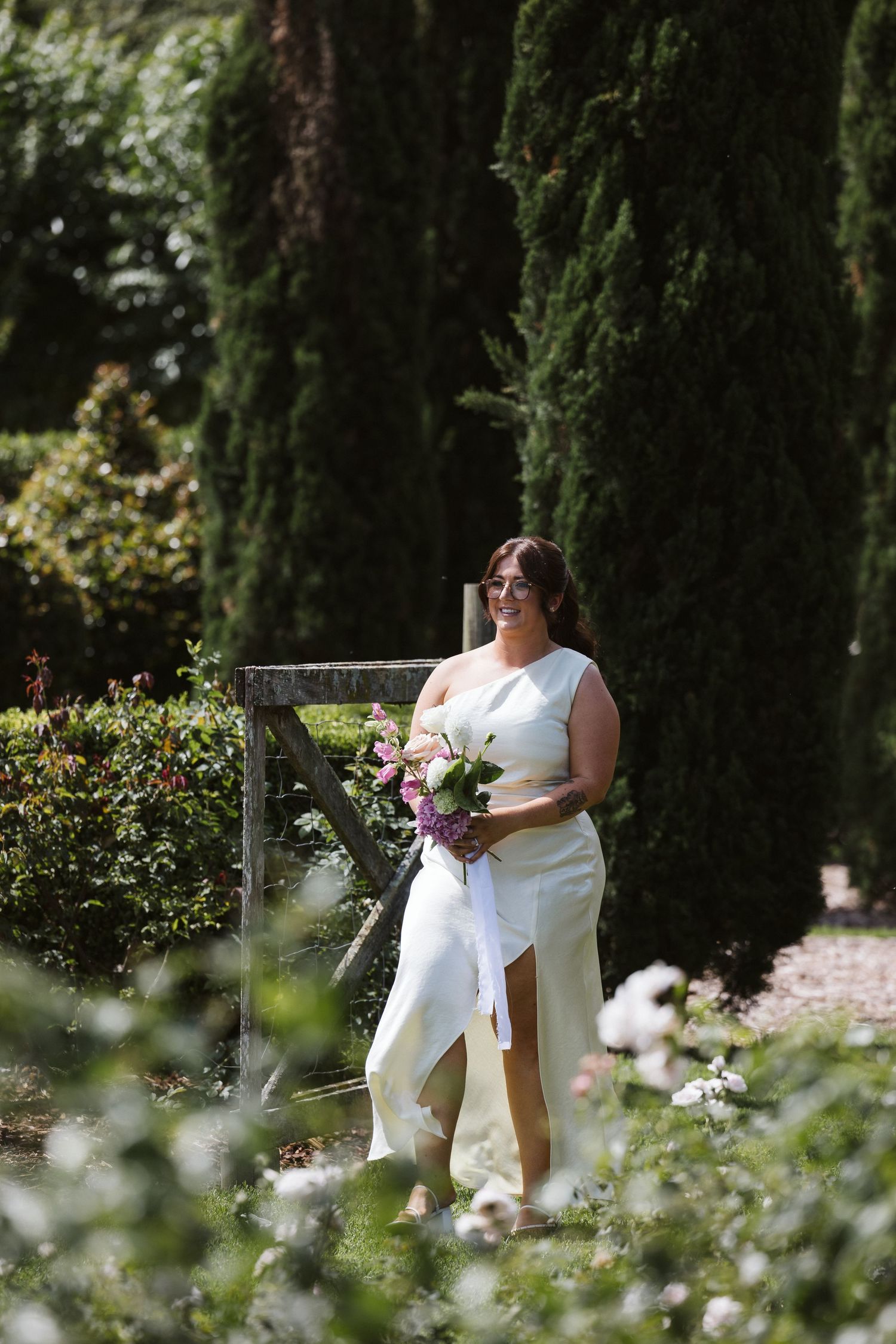 Person in white dress holds bouquet while standing in garden surrounded by tall cypress trees.