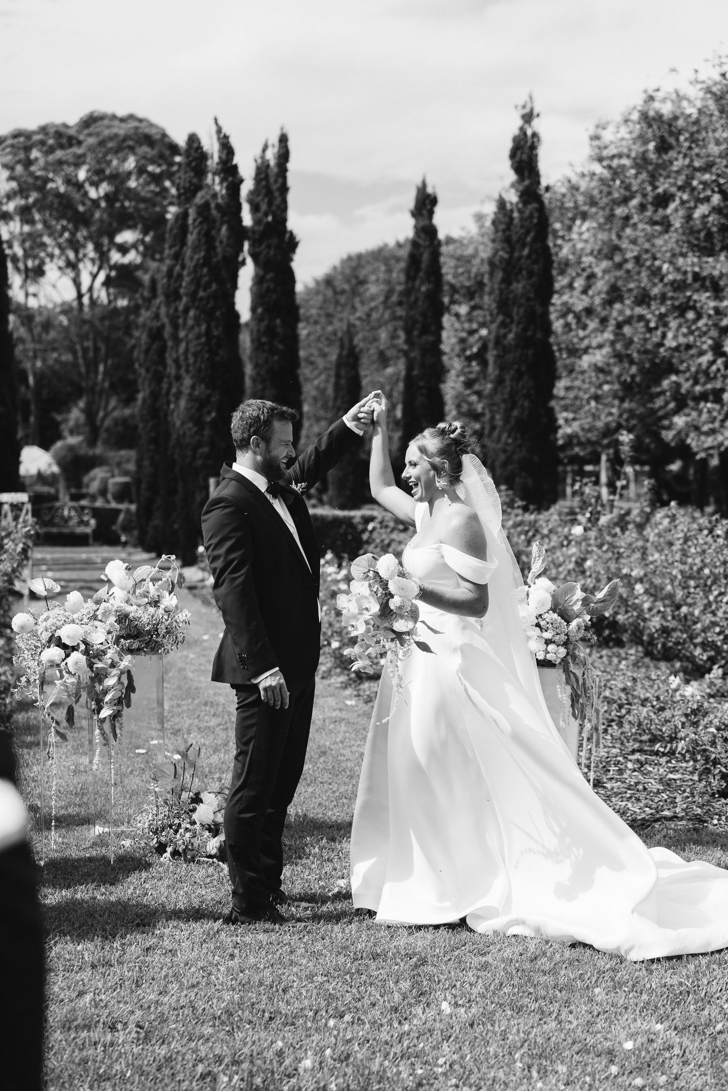 Black and white wedding portrait with tall cypress trees in garden background.