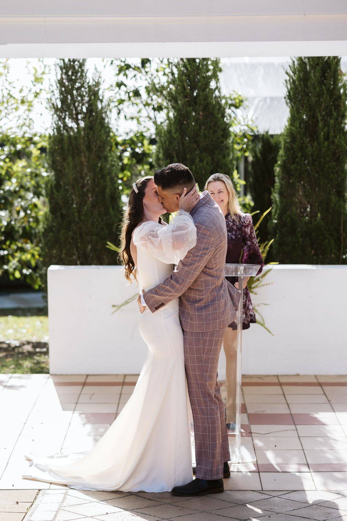 Bride and groom share first kiss at outdoor wedding ceremony with officiant and green trees in background.