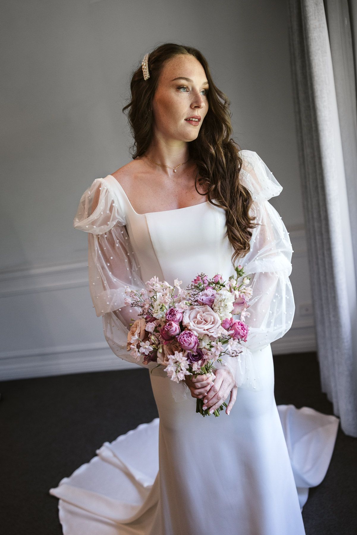 Bride in white gown with sheer puff sleeves holds pink floral bouquet, gazing softly to the side indoors.
