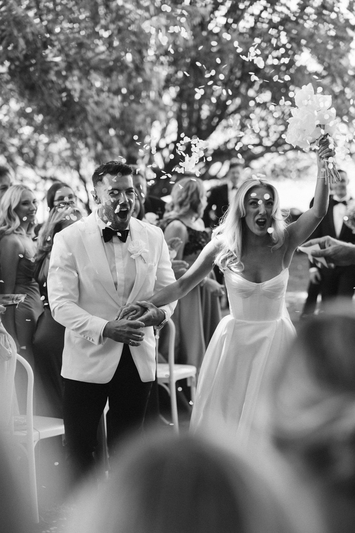 A black and white photo captures newlyweds celebrating with confetti at their outdoor wedding.