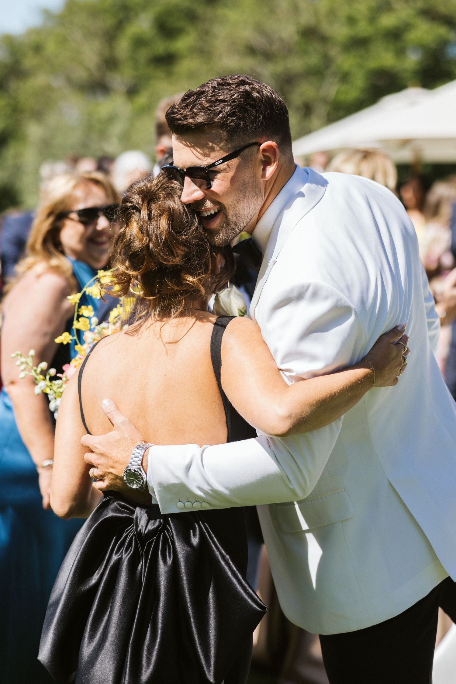 A warm embrace shared during an outdoor wedding reception with guests in background.