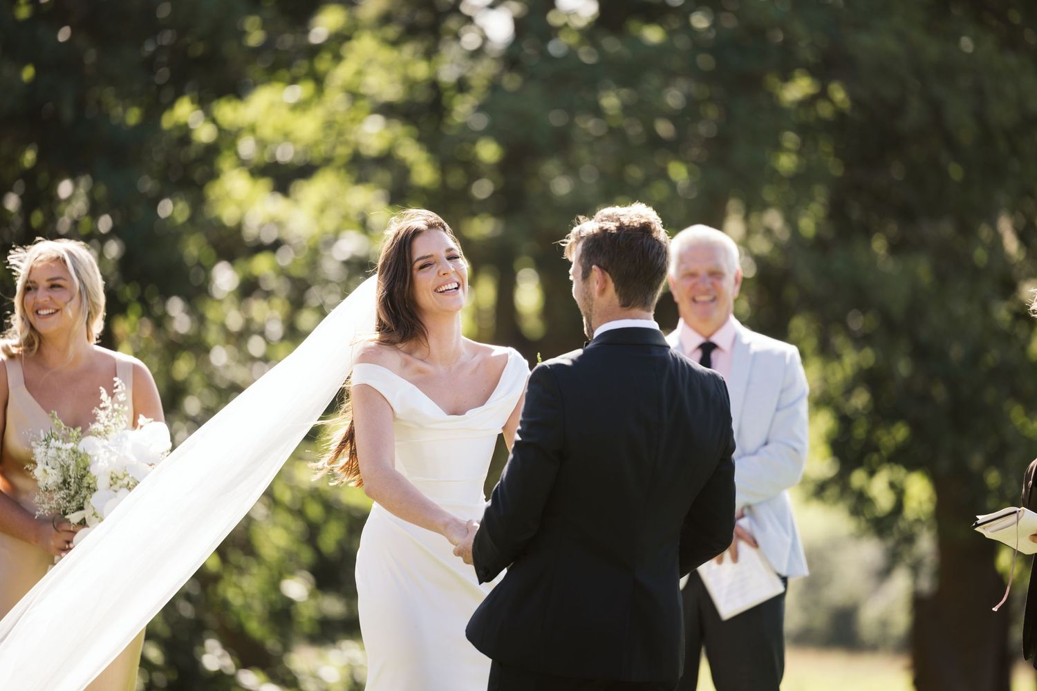Sunlit outdoor wedding ceremony with attendants and white floral decorations.