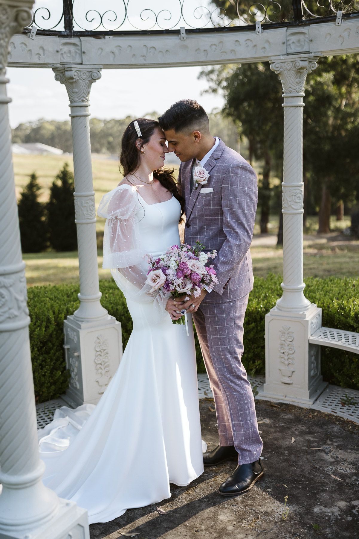 Bride and groom share an intimate moment under a white gazebo, she in a mermaid gown, he in a plaid suit.