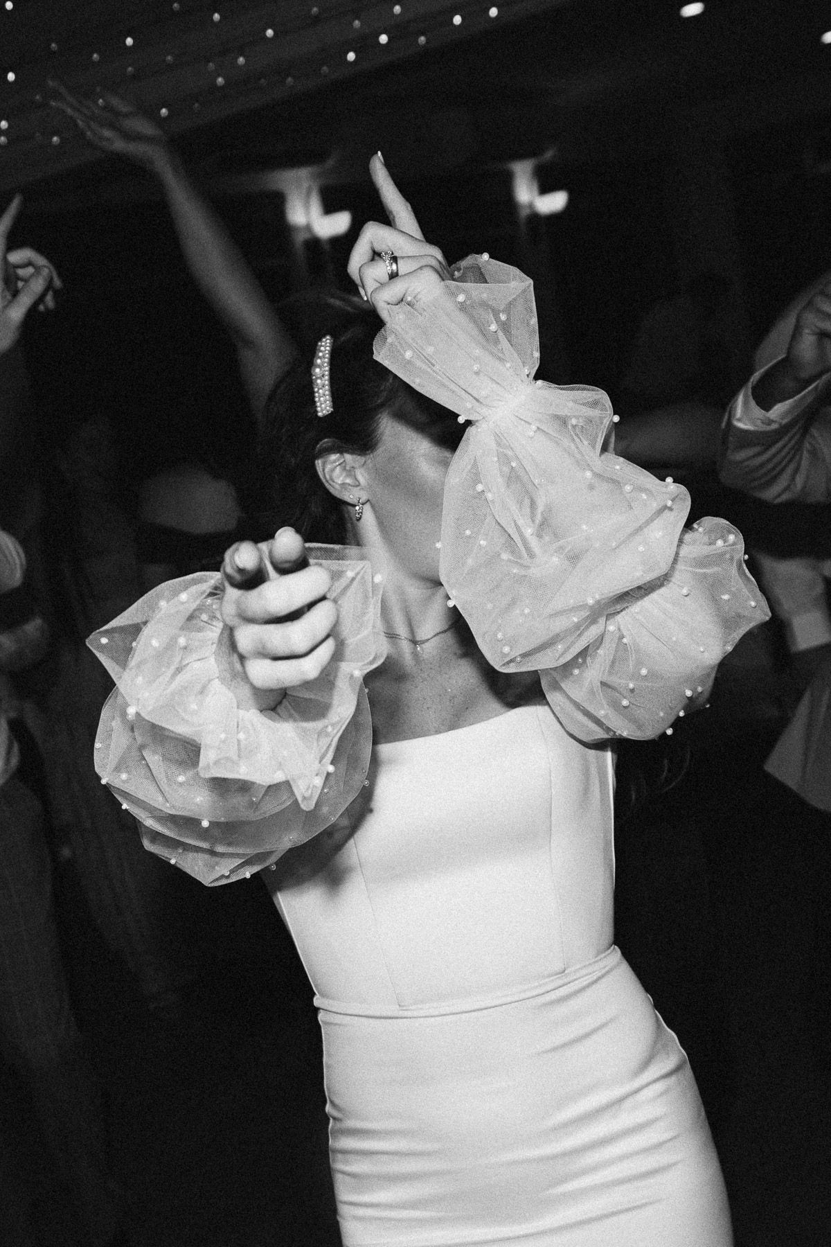 Bride in white dress with voluminous organza bow sleeves dancing joyfully at wedding reception, black and white photo.