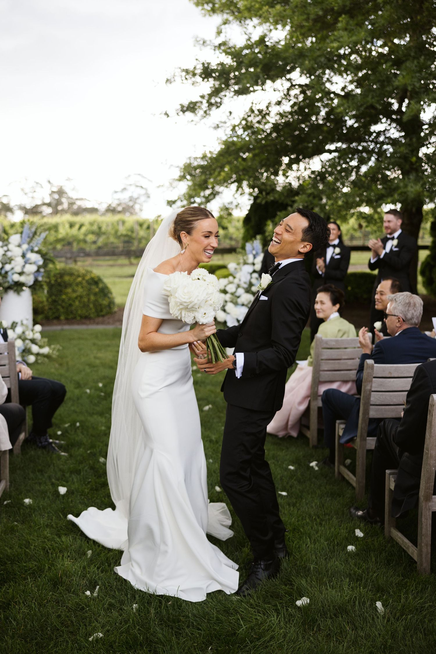Couple sharing a lighthearted moment during outdoor wedding ceremony with flower petals falling.