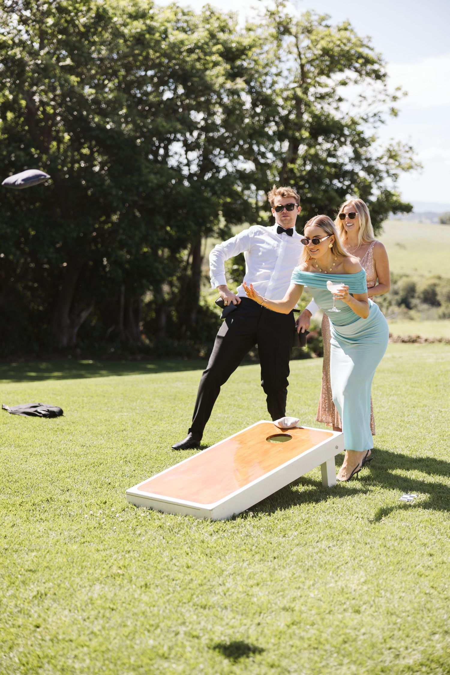 Wedding guests play cornhole on a sunny lawn during outdoor reception festivities.
