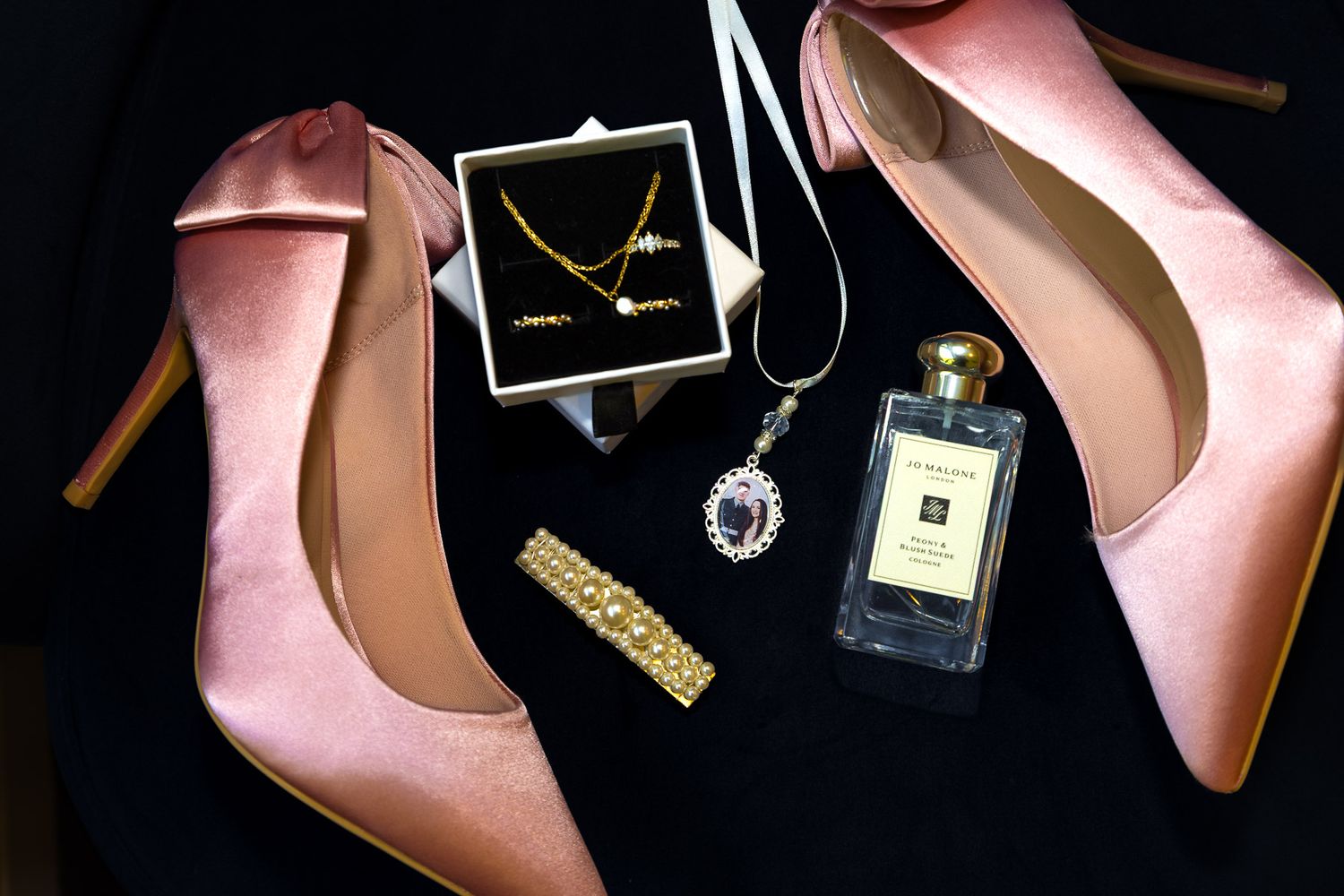Pink velvet heels displayed with gold jewelry, sapphire necklace, bracelet, and Jo Malone perfume on black background.