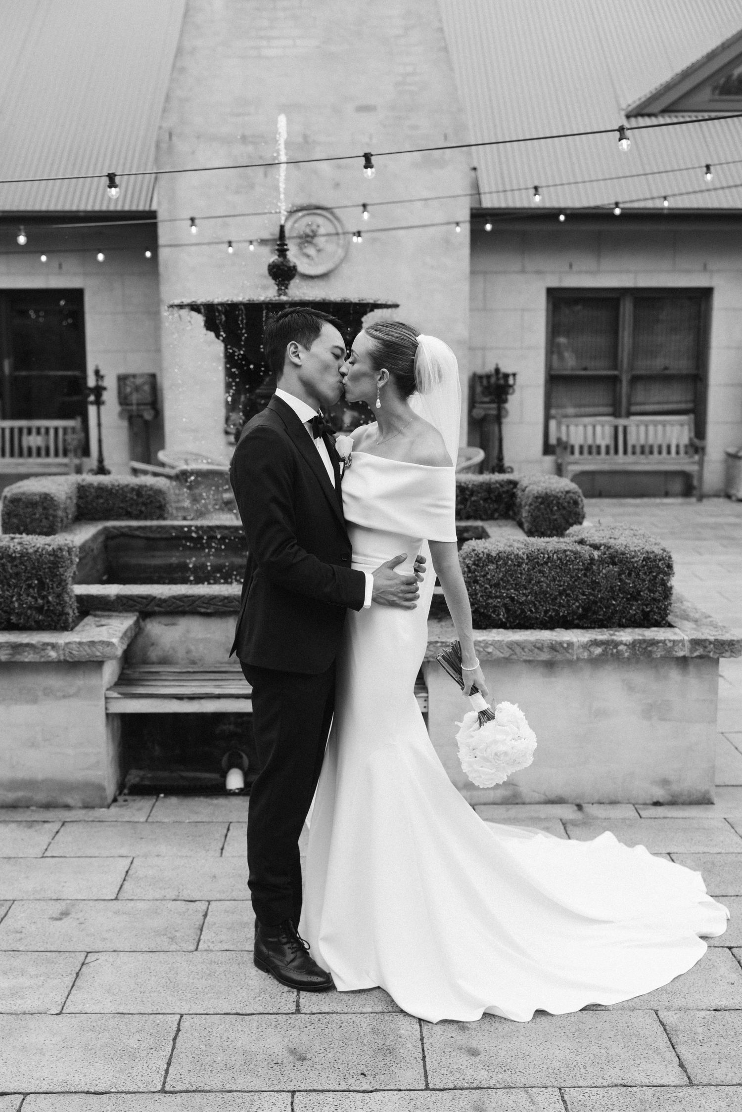 Black and white romantic portrait of bride and groom kissing in front of stone building with string lights.