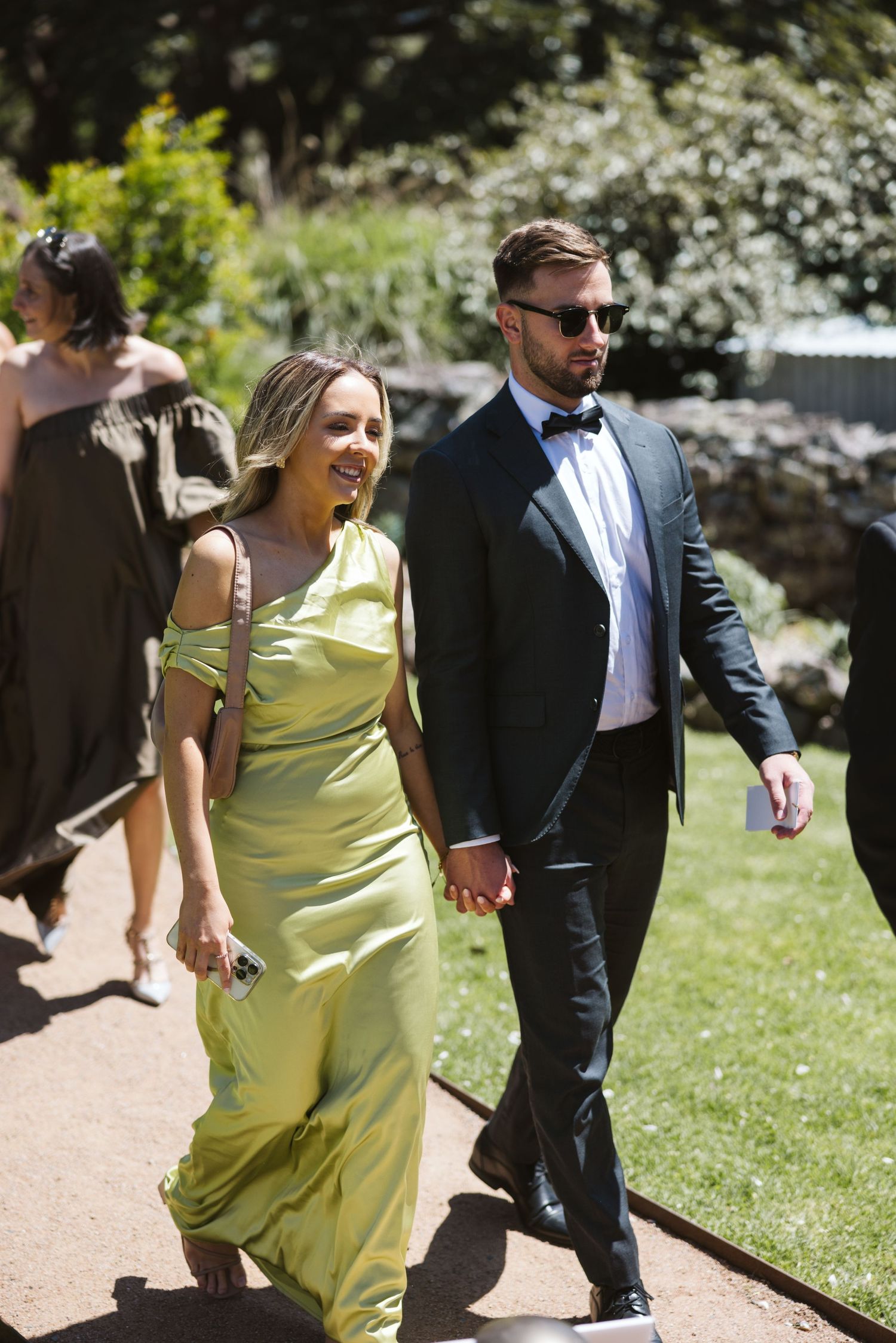 Couple in formal attire walking together on garden path, woman wearing yellow flowing dress.