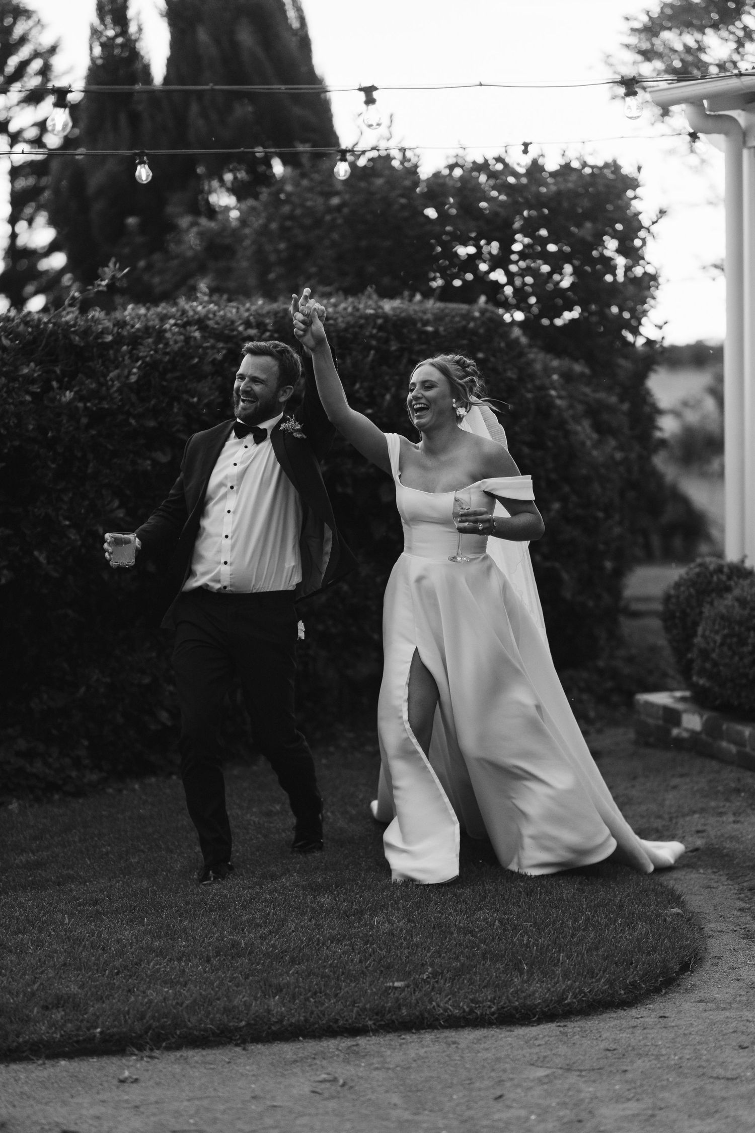 A joyful couple dances on a lawn at their wedding reception, with the bride wearing a flowing white gown with a slit.