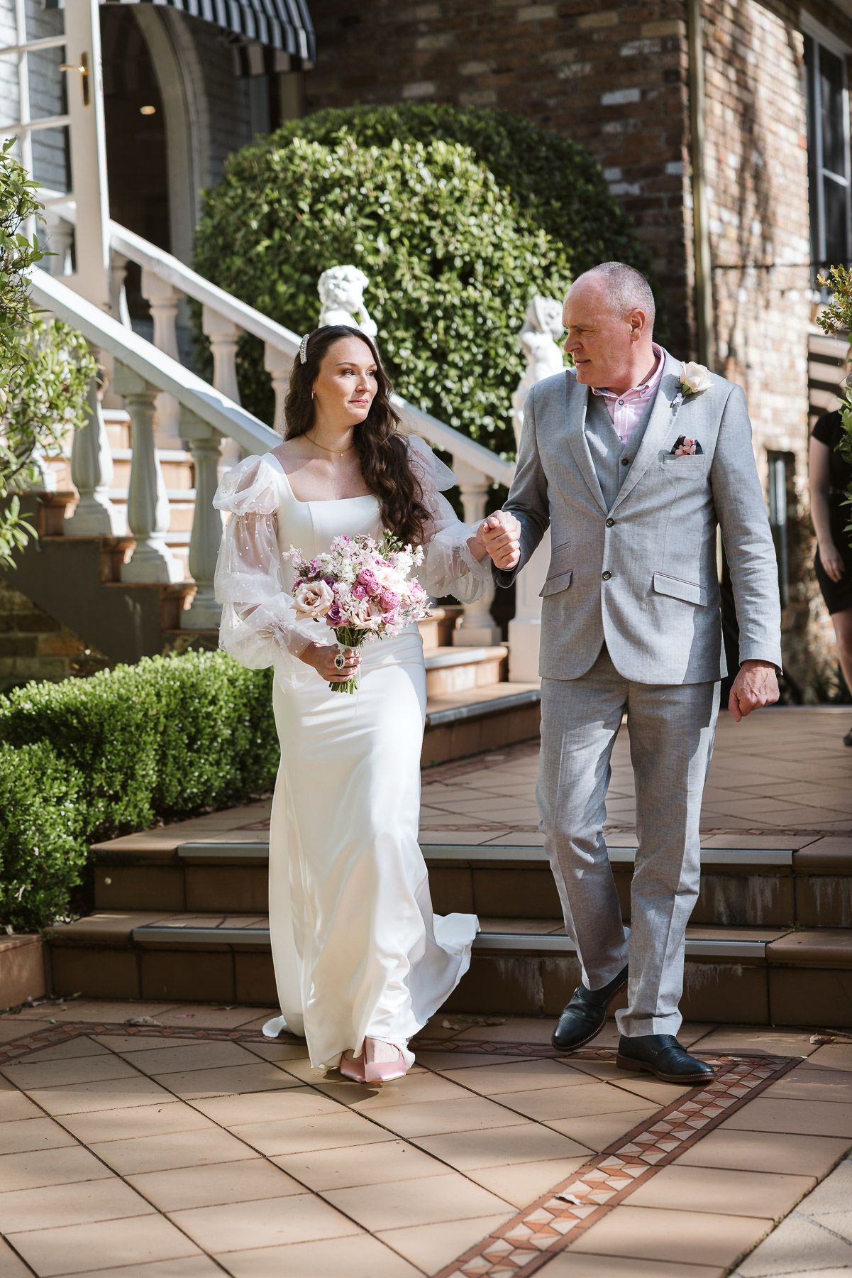 Bride in white gown holding pink bouquet walks hand-in-hand with man in gray suit down outdoor steps.