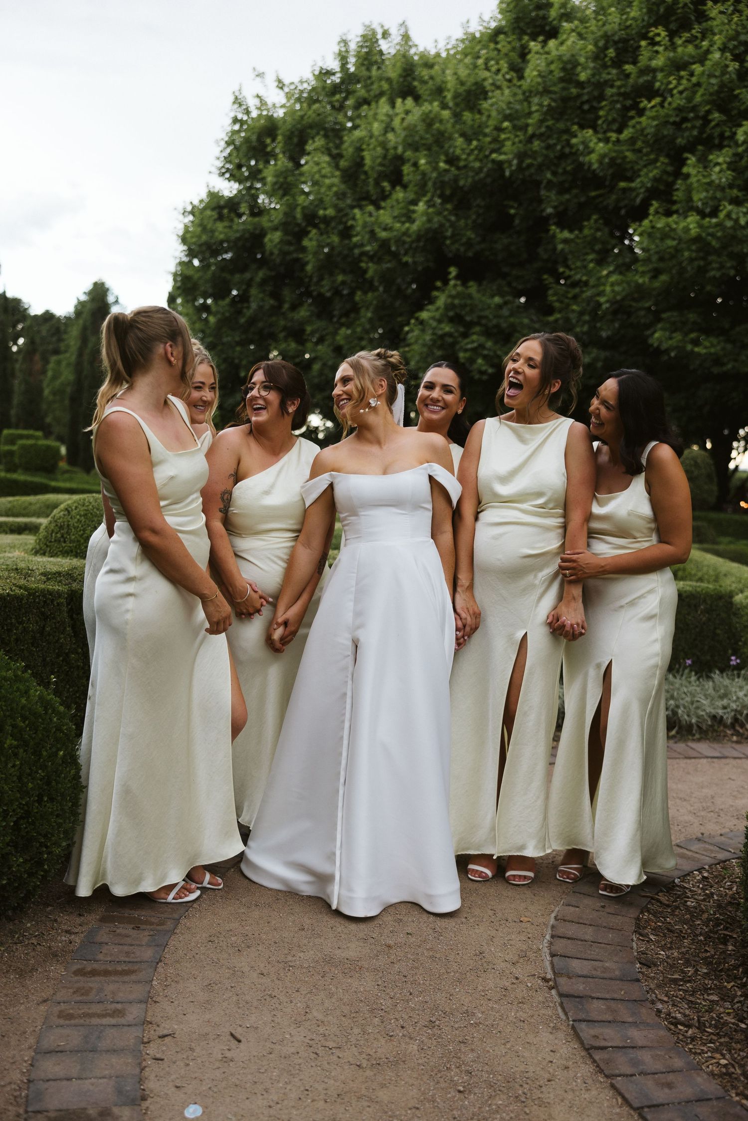 Bridal party in matching white dresses share a joyful moment on garden path.