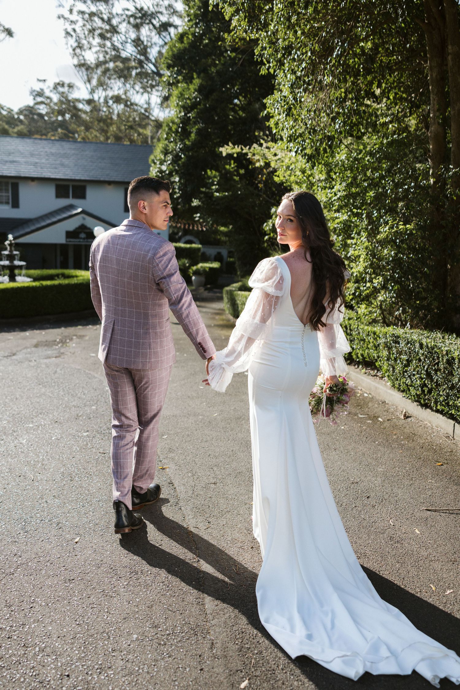 A newlywed couple walking hand in hand on a tree-lined path, bride in white and groom in lavender suit.