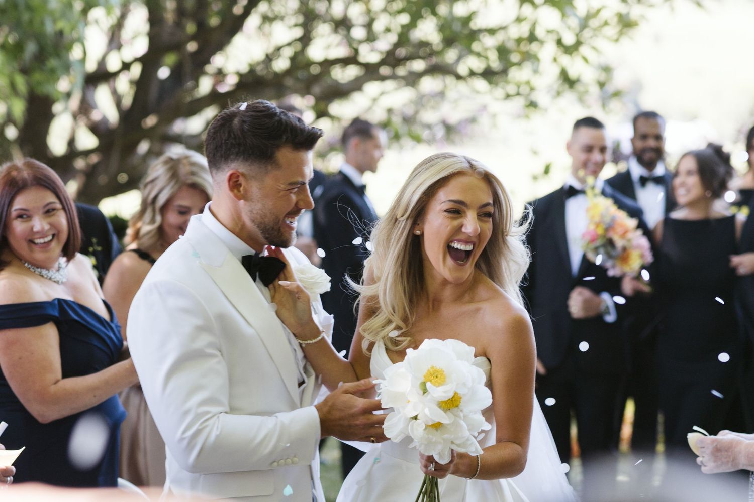 Newlyweds share a laugh while holding a white bouquet during their outdoor wedding celebration.
