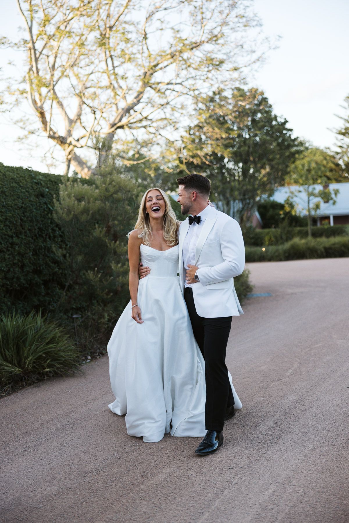Bride in white gown and groom in white tuxedo laughing together on a garden path at their wedding.