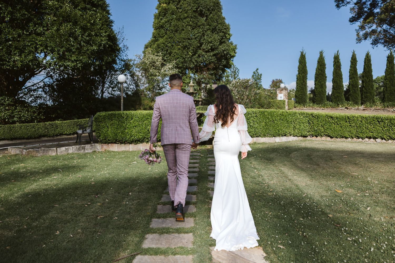 A bride and groom walk hand in hand through a lush green garden on their wedding day.