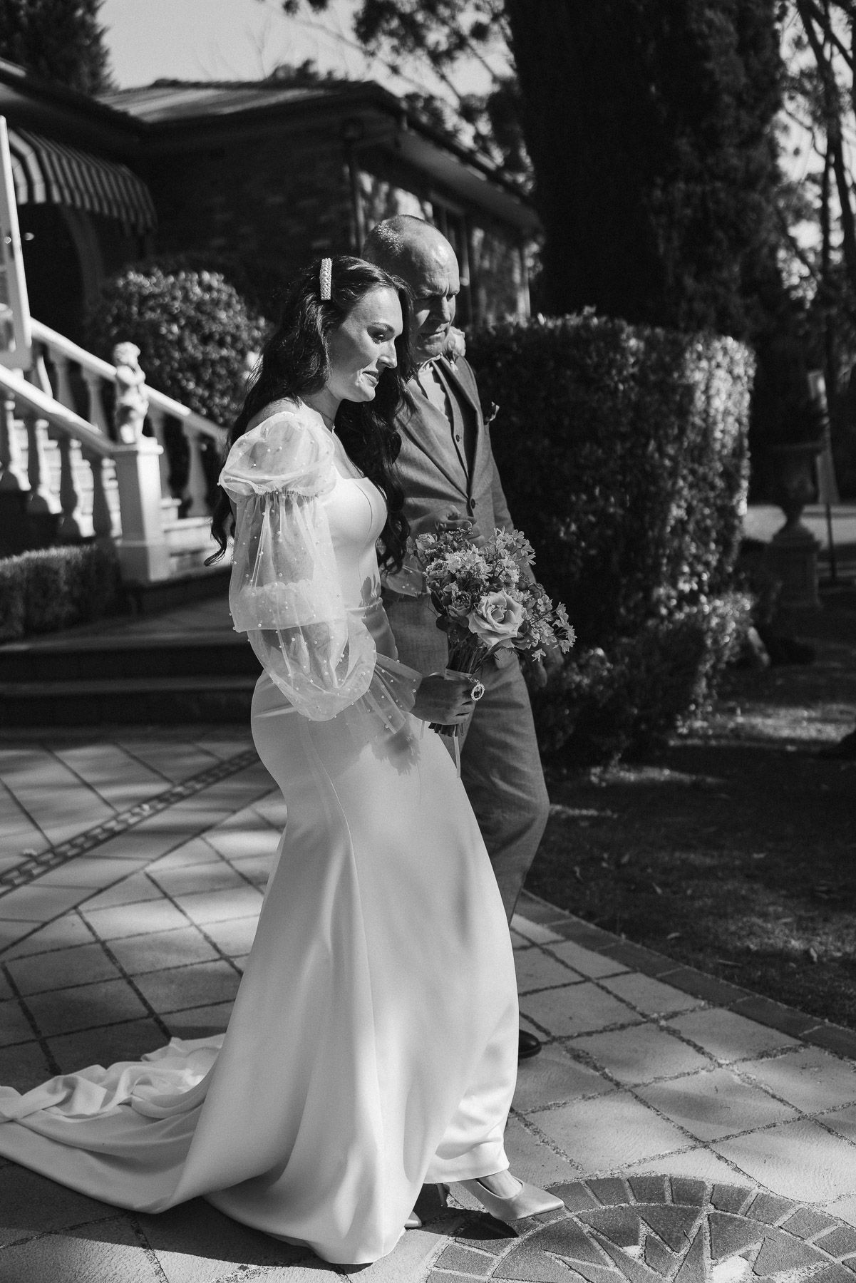Black and white photo of a bride in a white gown with sheer sleeves walking with a man in a suit outdoors.