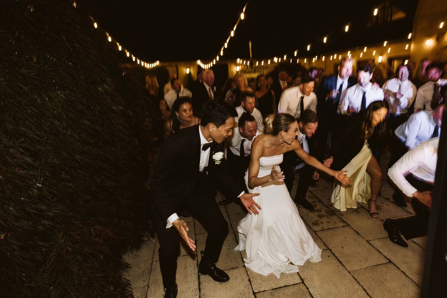Wedding guests dancing and celebrating on a stone patio illuminated by string lights.