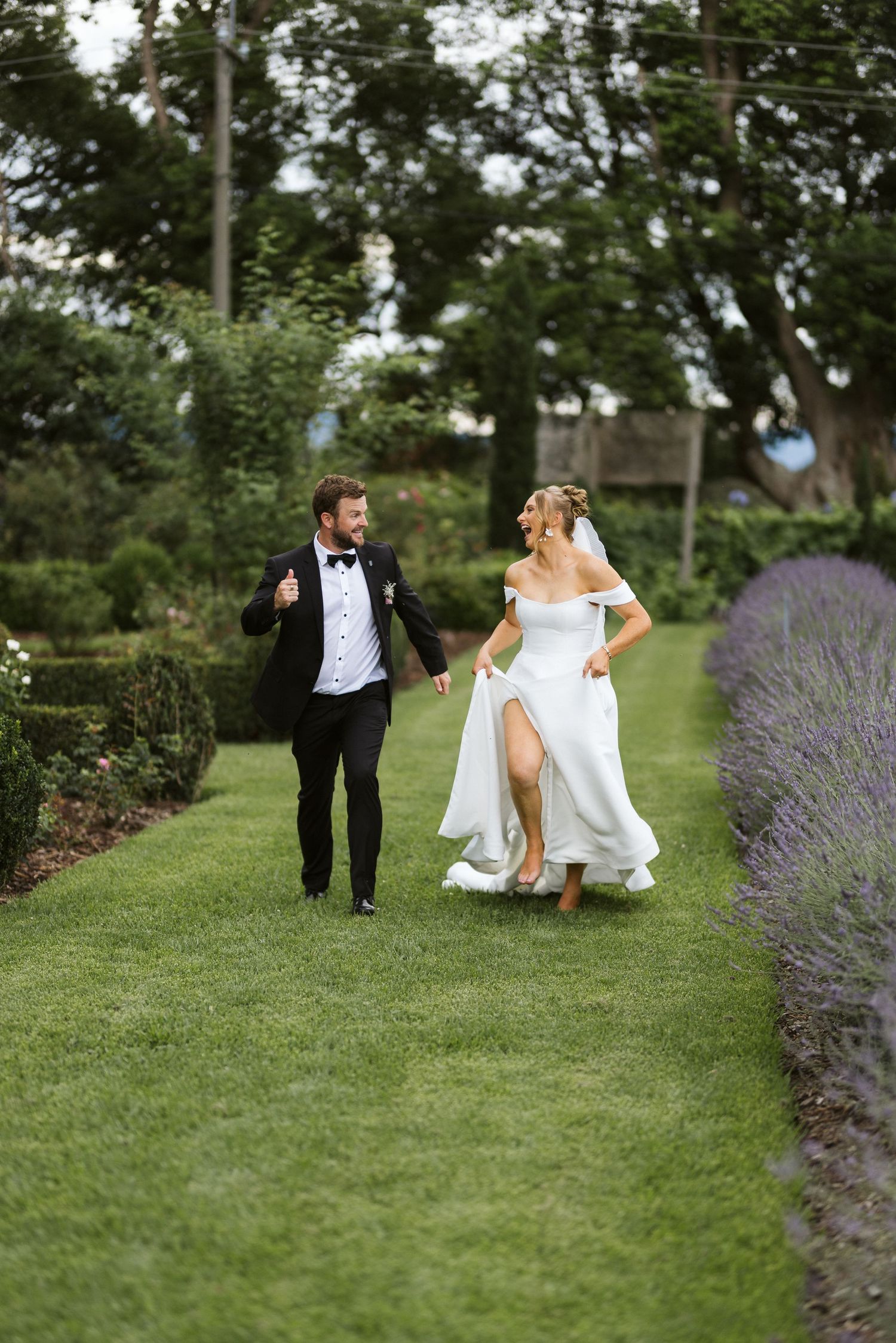 Playful couple runs along garden path lined with lavender during wedding celebration.