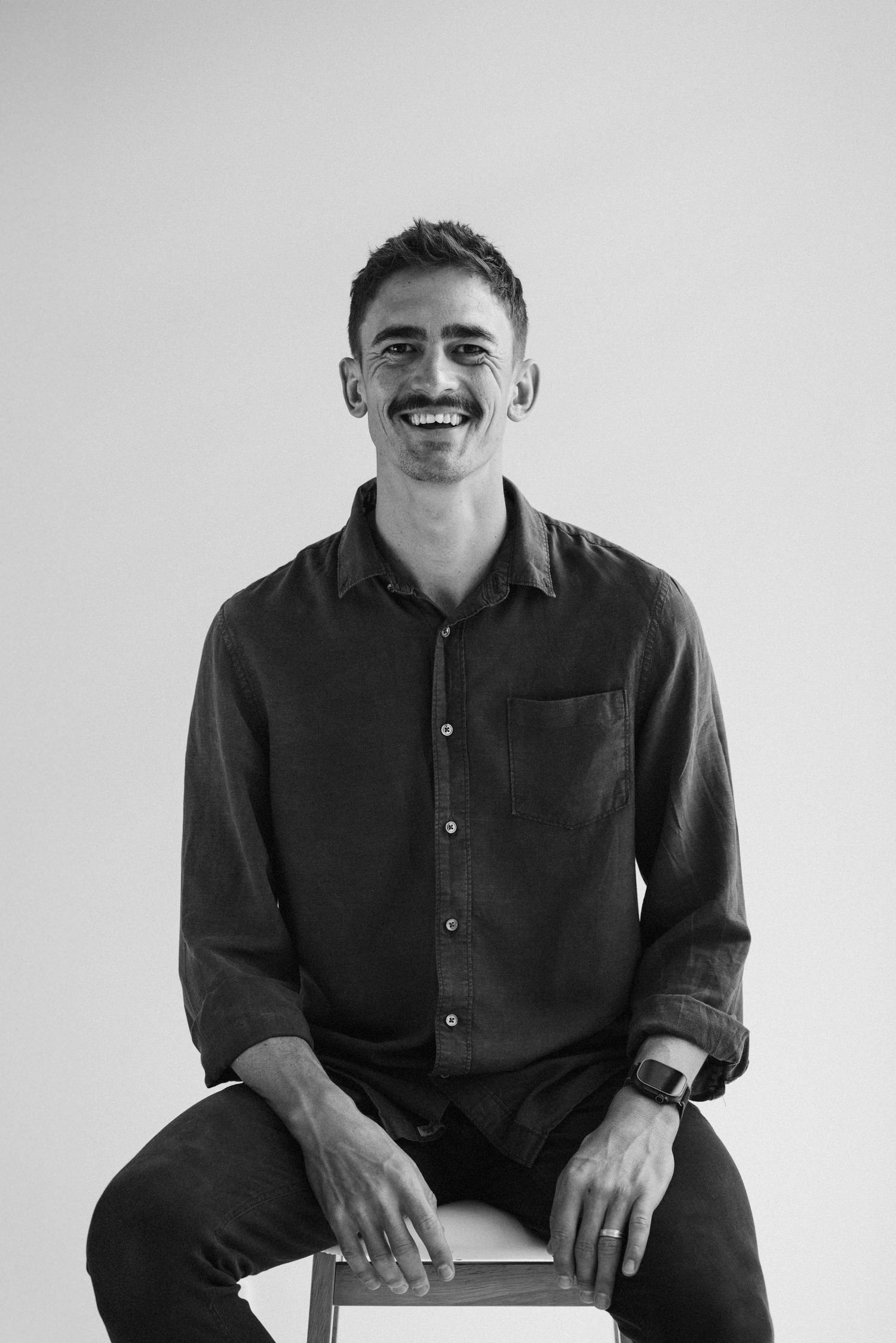 A person wearing a dark button-down shirt sits casually on a chair against a white background.