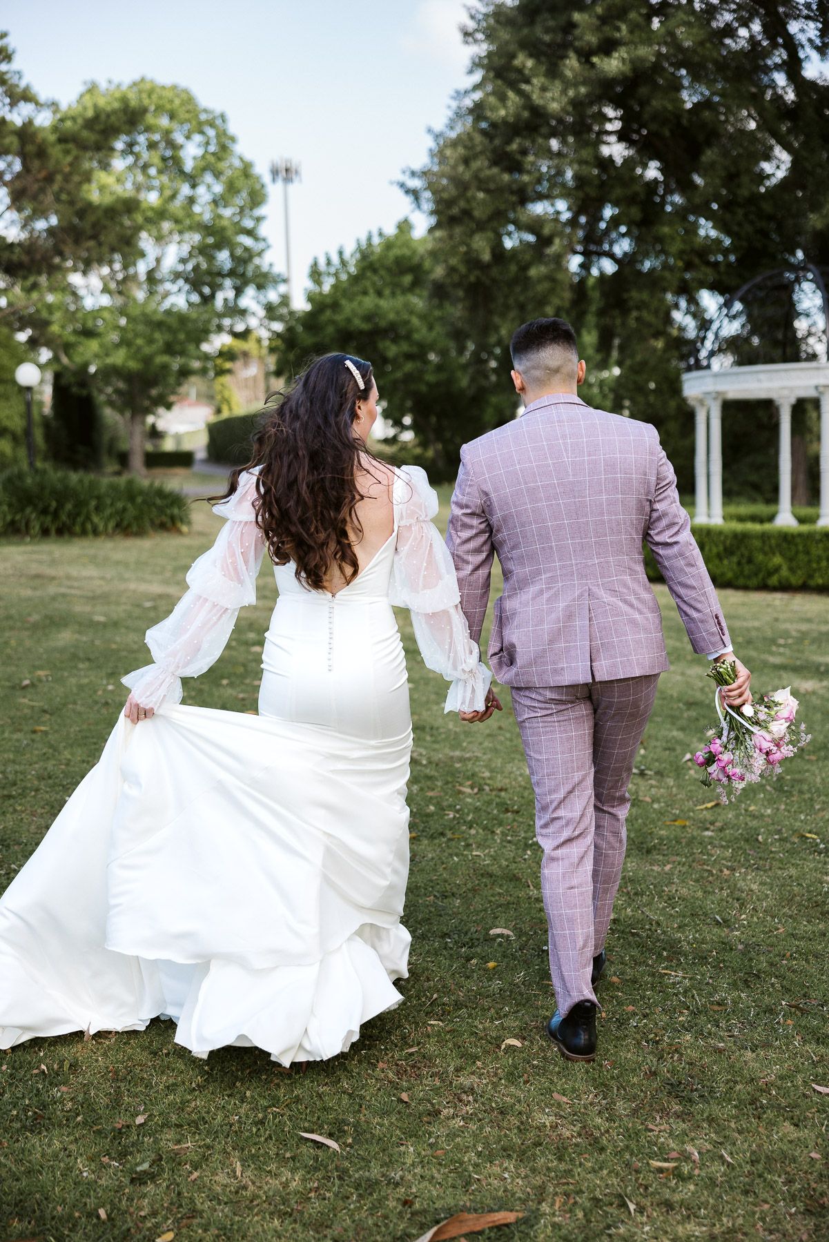 Bride in white gown and groom in plaid suit walk hand-in-hand through lush green park on wedding day.