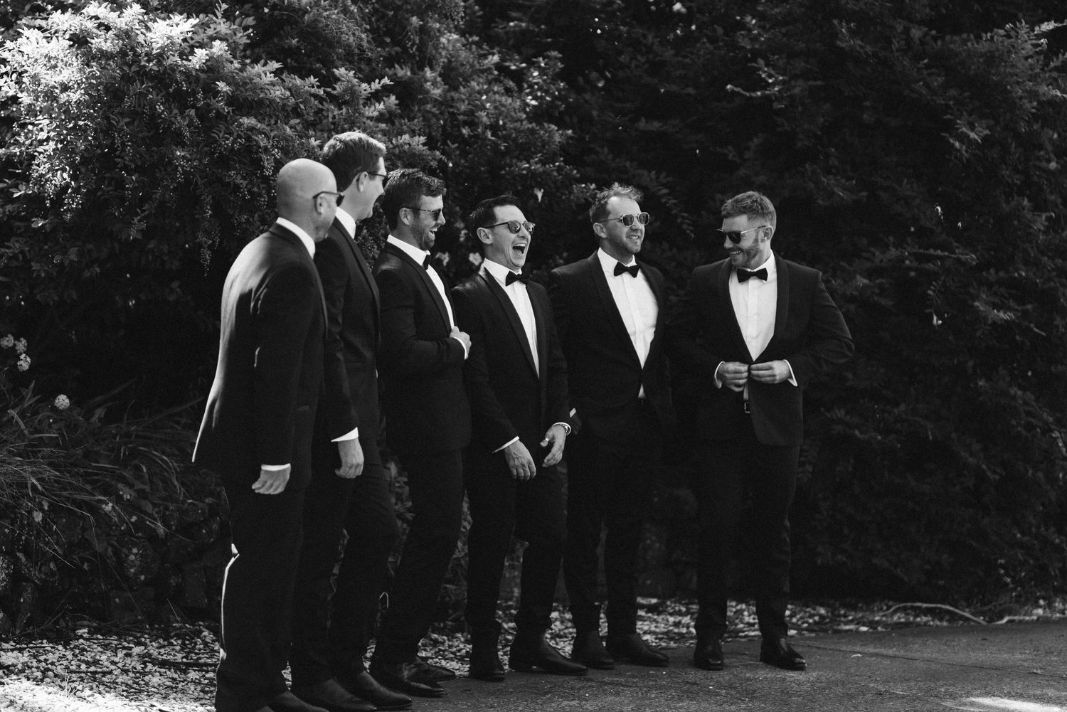 Group of groomsmen in black tuxedos standing together outdoors in black and white.
