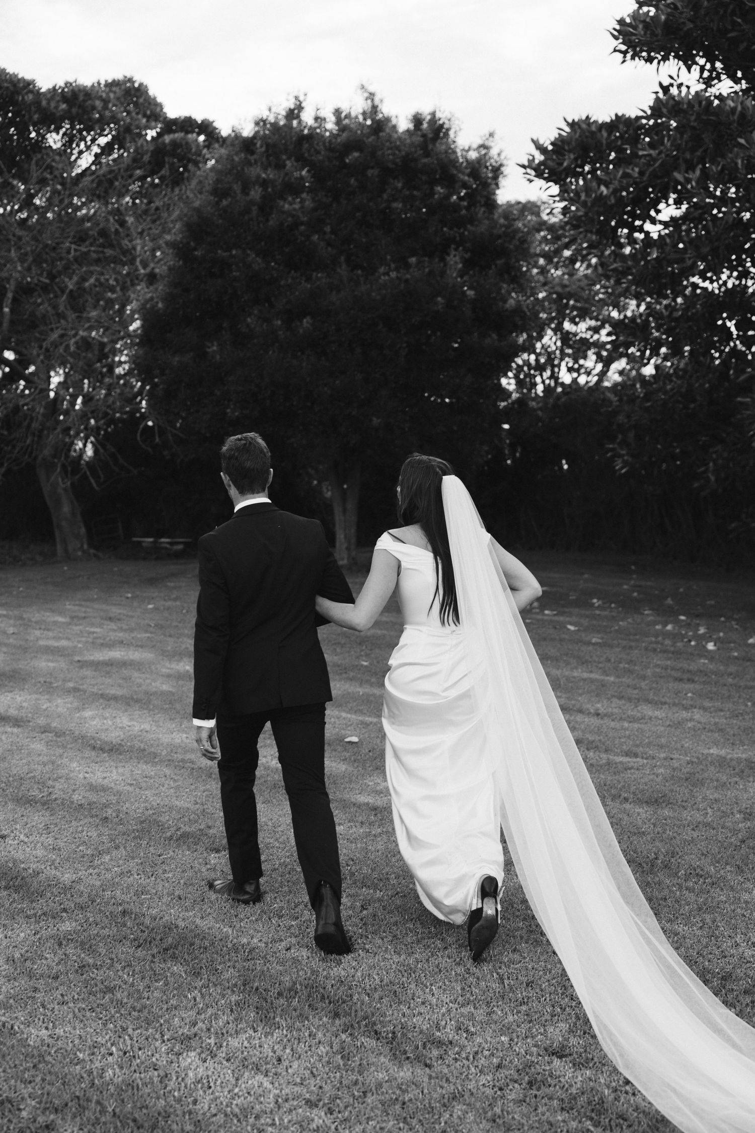 Black and white photo of newlyweds walking away with bride's veil trailing behind on grassy lawn.