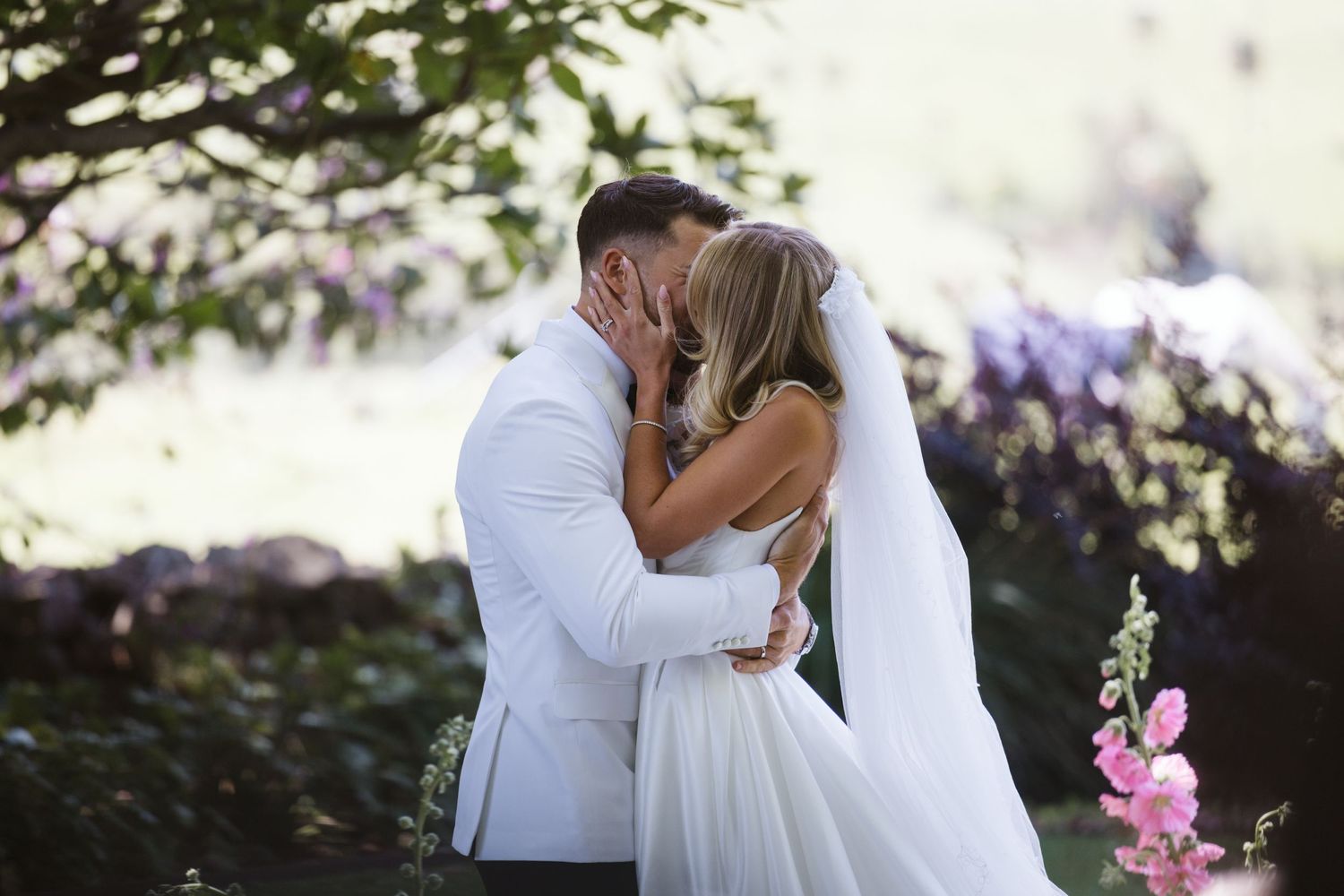 Romantic kiss shared during wedding ceremony with pink flowers in background.