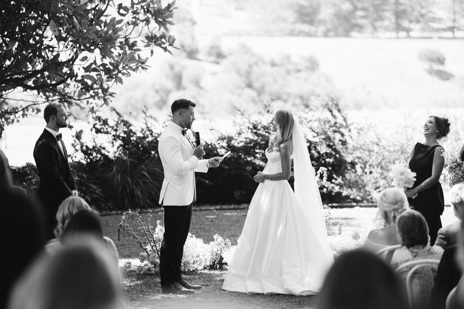 Black and white photo of a wedding ceremony taking place under trees with guests seated.