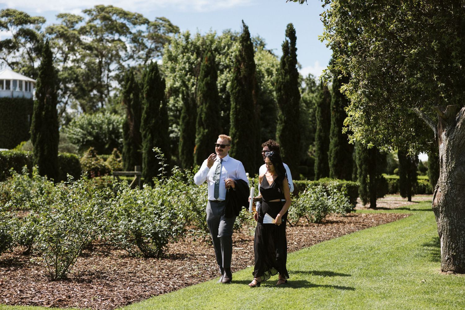 Well-dressed couple strolling along landscaped garden path with cypress trees.