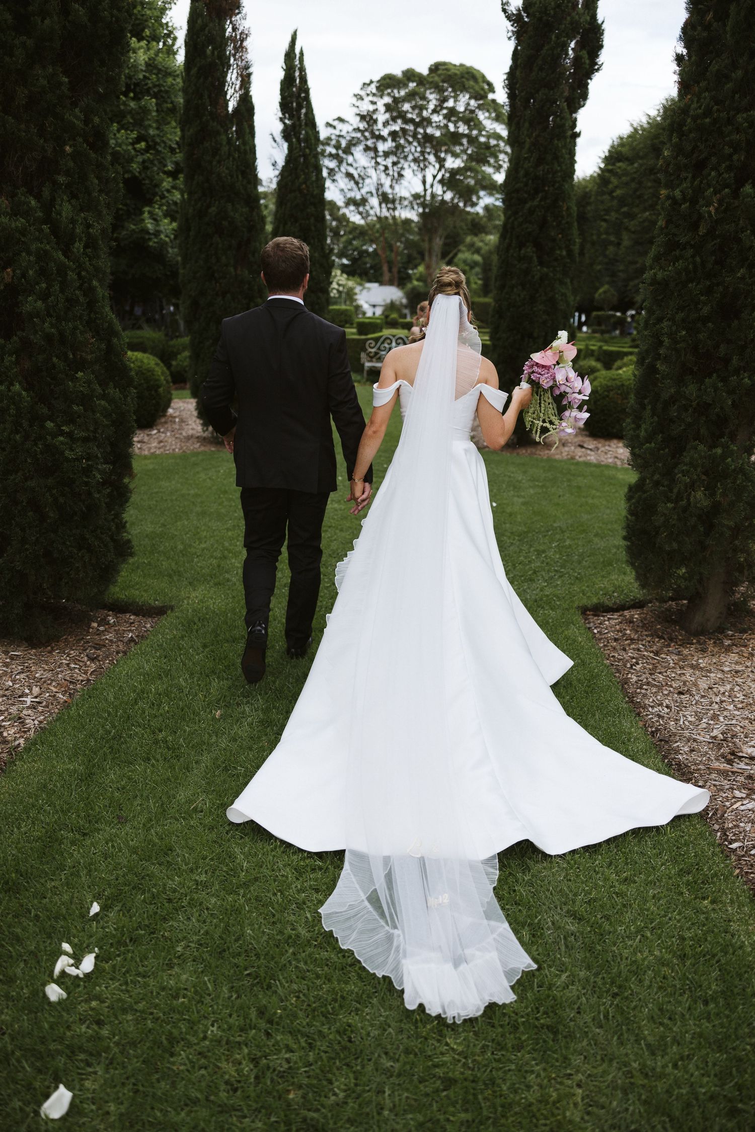 Newlyweds walk down a garden path lined with tall cypress trees, bride's white dress trailing behind.