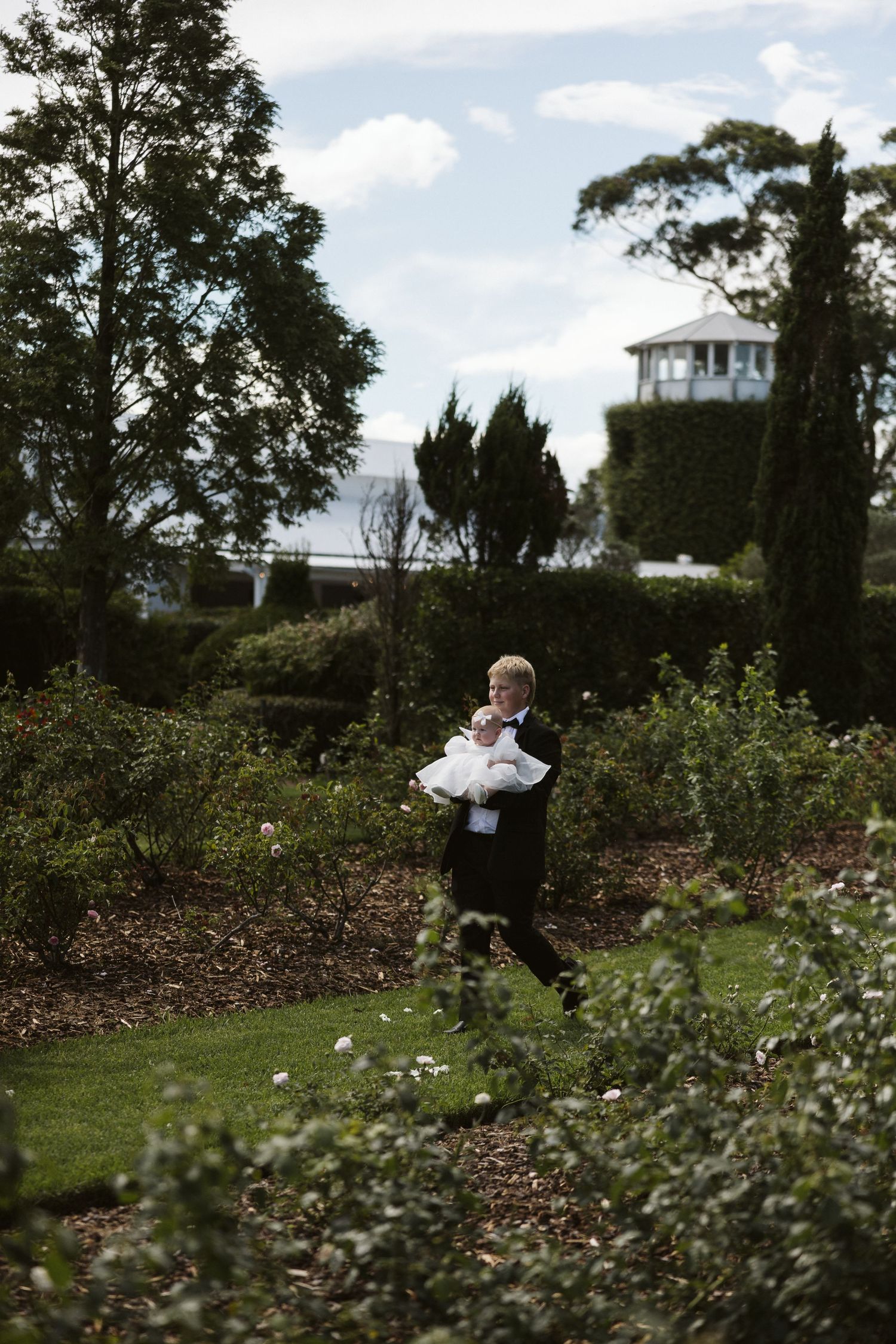 Small attendant in formal wear walks through a lush garden with a white house visible in the background.