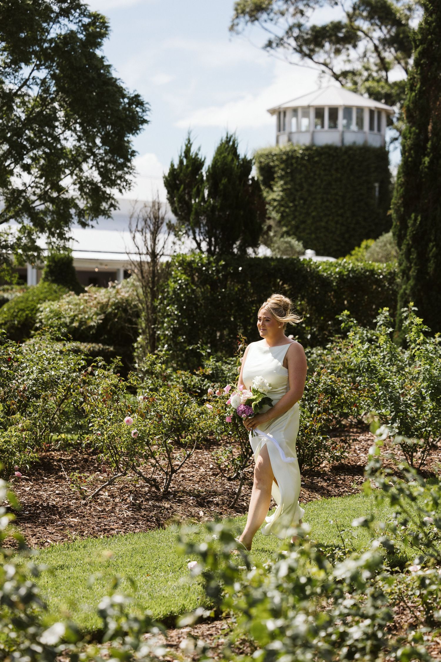 Person in elegant white dress walks through garden path with white lighthouse-style building in background.