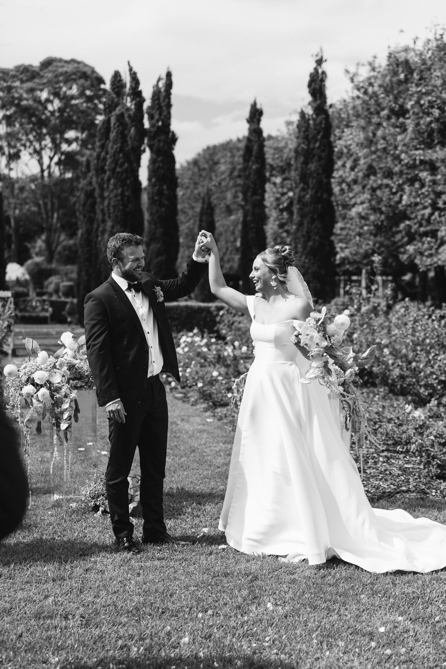 Black and white wedding photo capturing a celebratory moment in a formal garden setting.
