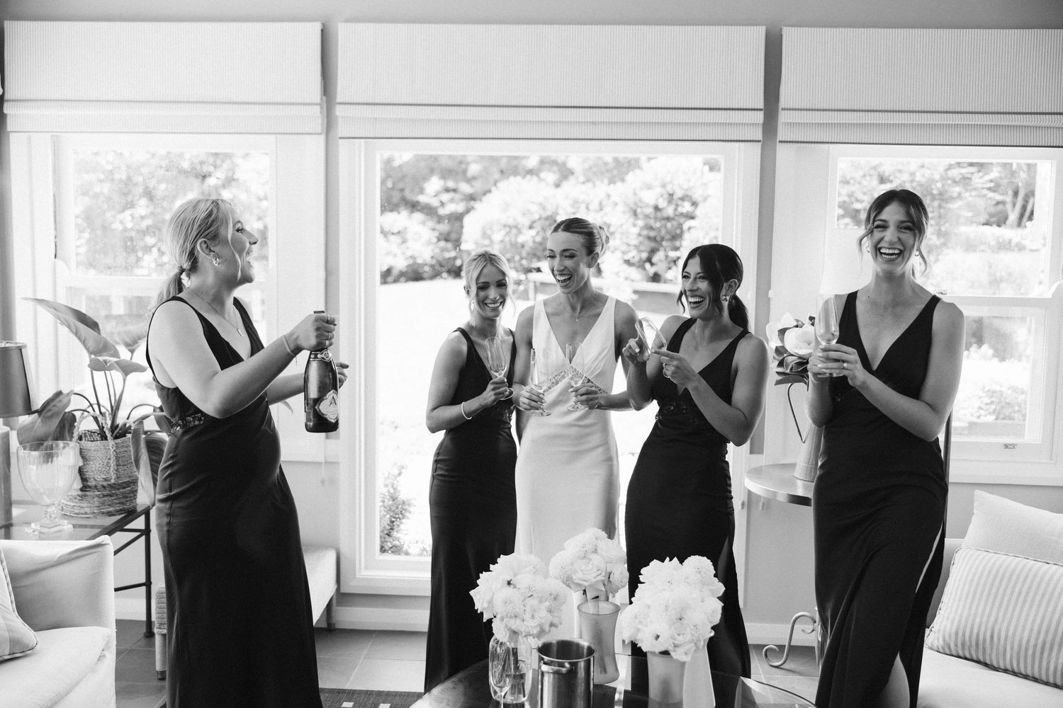 Black and white photo of bridesmaids celebrating with champagne in a sunlit room.