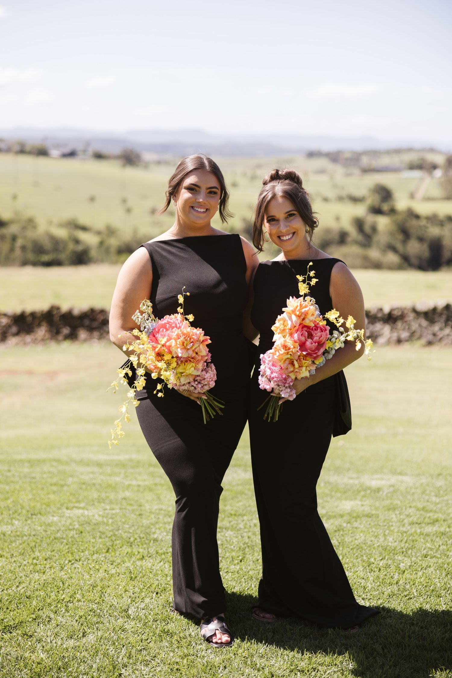 Two bridesmaids in black dresses hold colorful bouquets while posing on a grassy hillside.