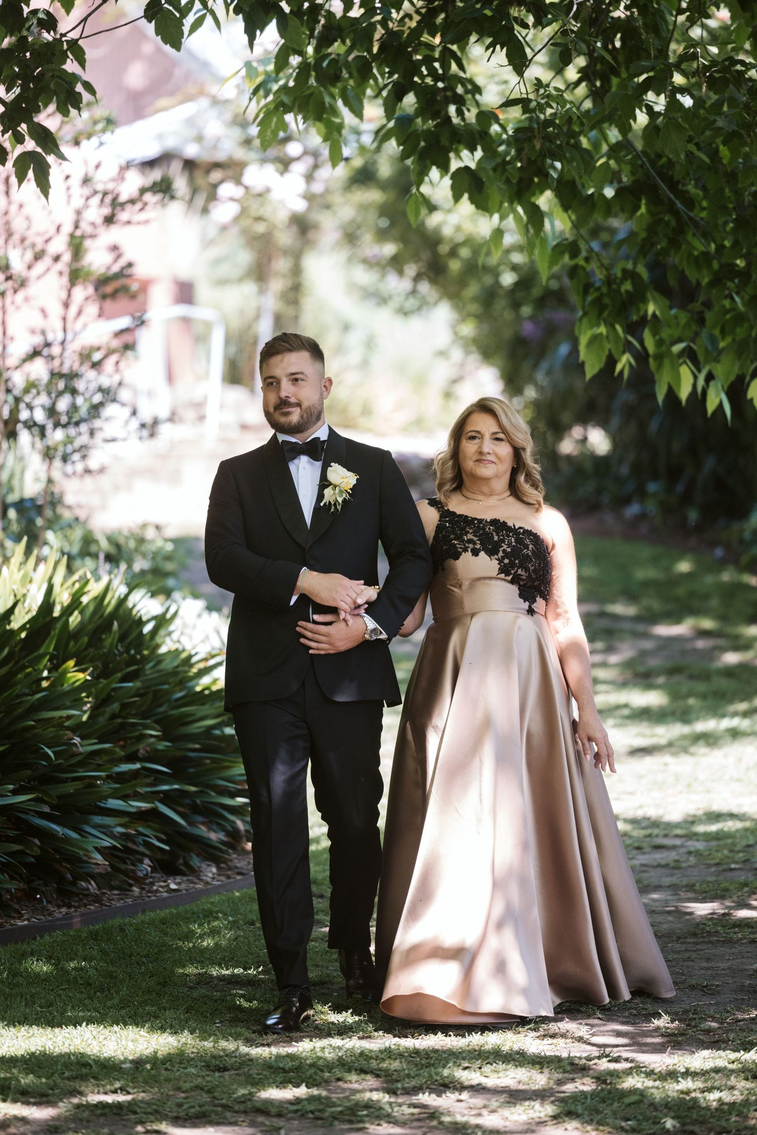 Formal couple walking arm in arm down garden path at elegant event.