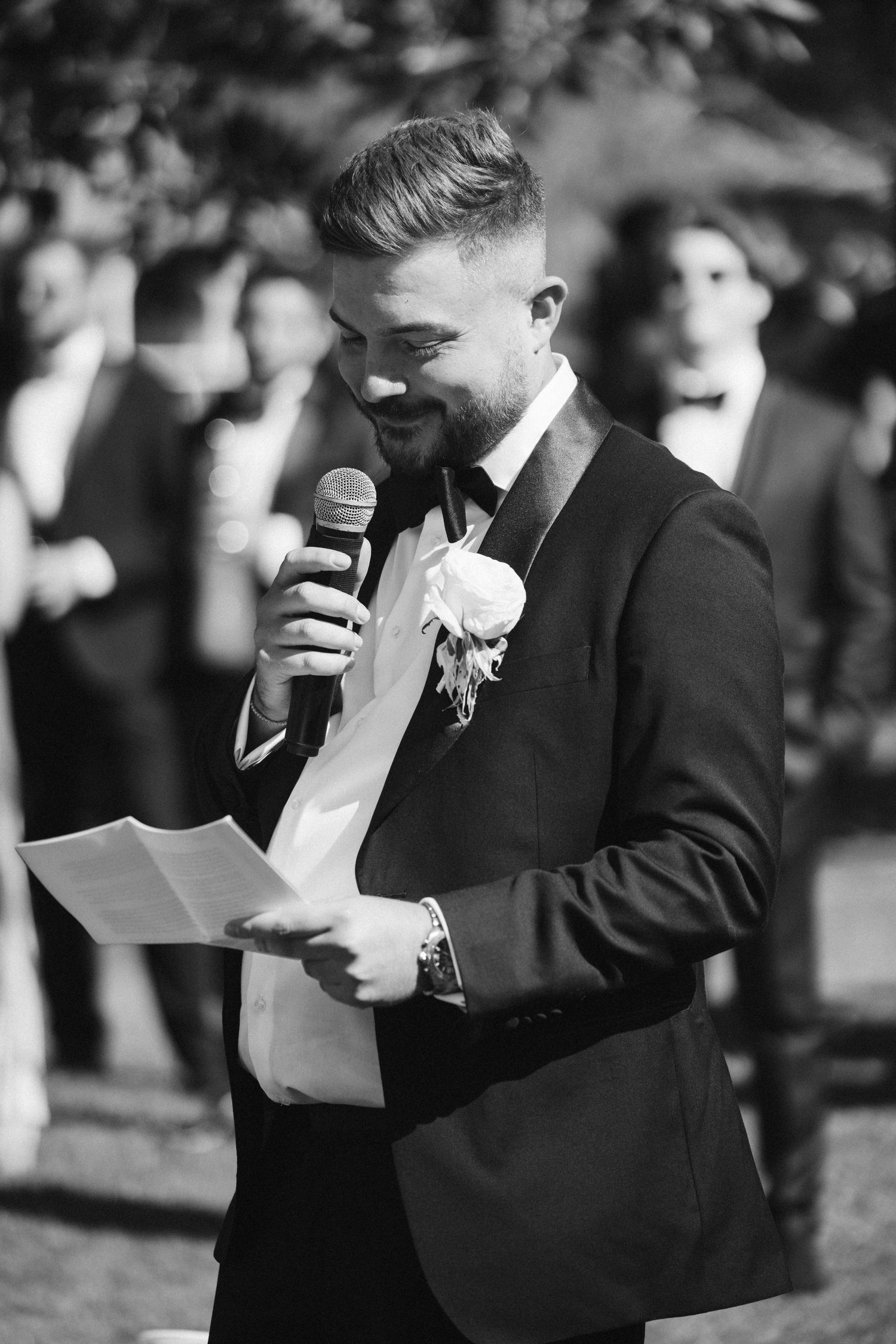 Wedding speech being delivered outdoors in classic black formal attire with boutonniere.