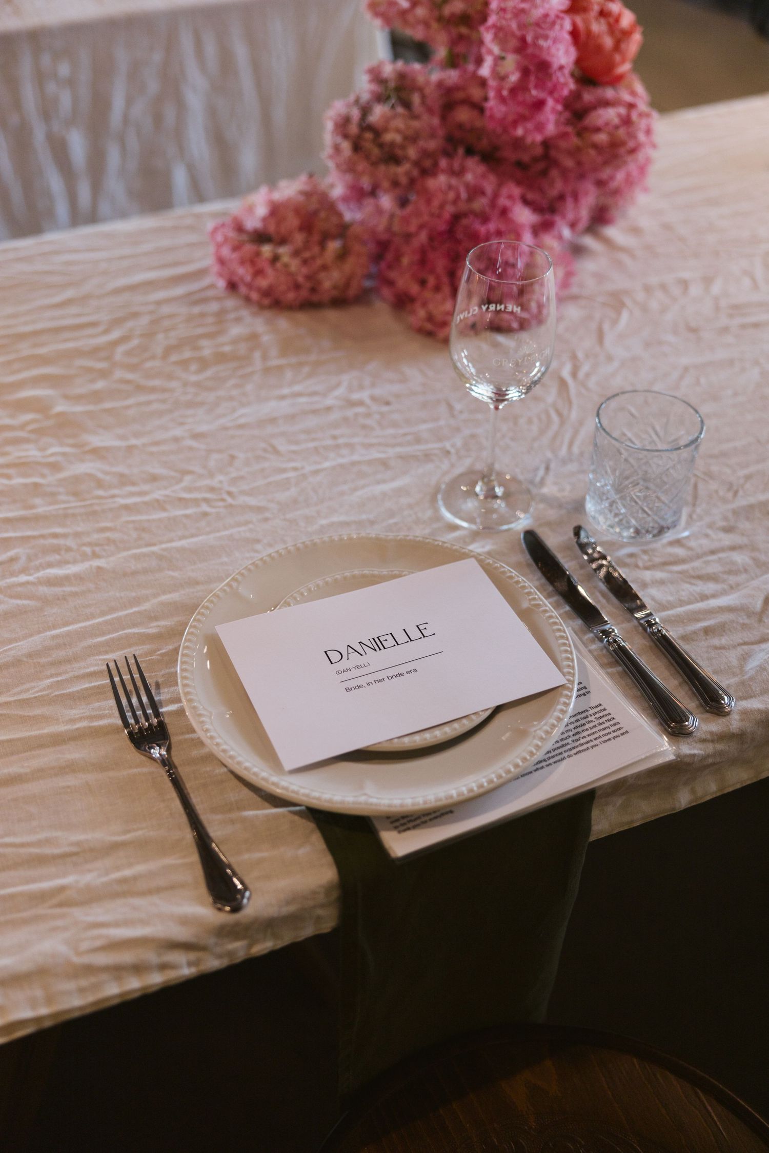 Place setting with name card, elegant glassware, and pink flowers on textured white tablecloth.