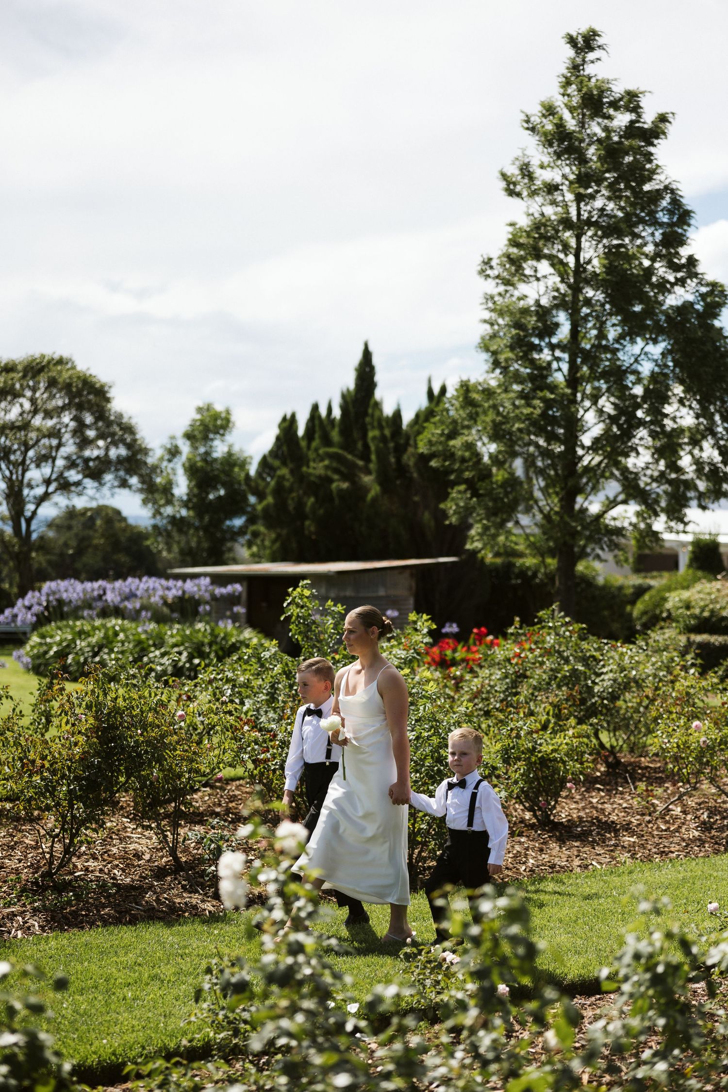 Wedding party walks through a beautiful garden with blooming flowers and tall trees in the background.