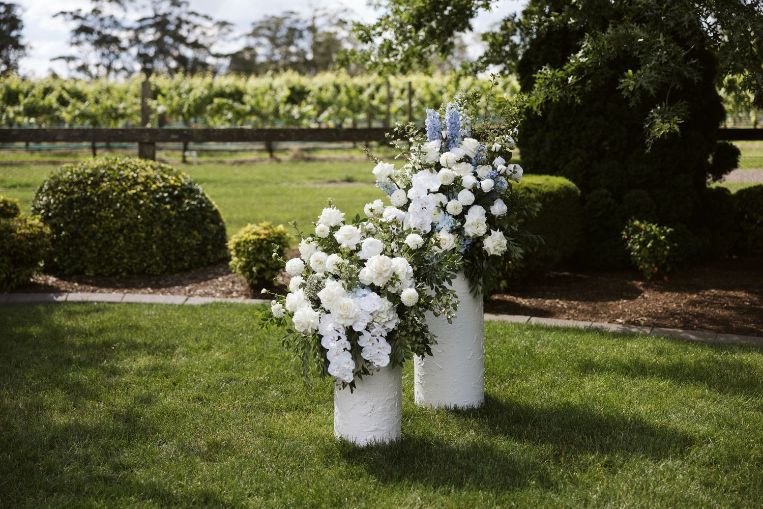 White floral arrangements on pedestals decorating an outdoor garden wedding space.