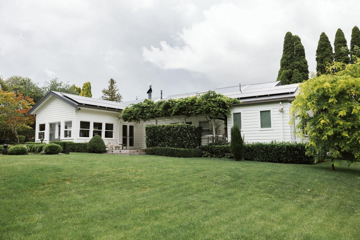 A white residential house with green lawn and cypress trees under cloudy skies.